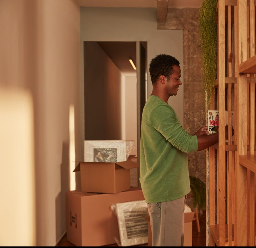 Homem organizando a estante da casa, com caixas de papelão de mudança ao fundo