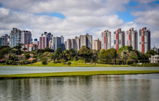 Foto que ilustra matéria sobre qualidade de vida em Curitiba mostra uma panorâmica do Parque Barigui, com um espelho d’água em primeiro plano, árvores no meio da imagem e altos prédios ao fundo