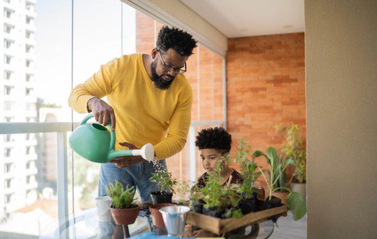 Foto que ilustra matéria sobre Como fazer horta em casa mostra um homem negro, de camisa amarela comprida, usando um regador para molhar pequenos vasos de plantas com ervas ao lado de seu filho em uma sacada de apartamento