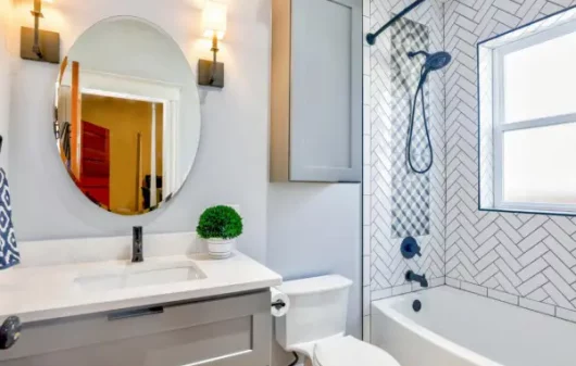 Banheiro pequeno minimalista em tons de branco, preto e cinza com espelho redondo, banheira, luminárias de parede e móveis planejados.