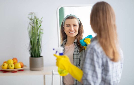Foto que ilustra matéria com dicas sobre como limpar espelho mostram uma mulher de costas, posicionada de frente para um espelho. Ela aparece sorrindo no reflexo, com um luvas amarelas, com um pano verde esfregando o espelho em uma das mãos e um borrifador na outra.