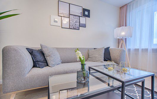 A foto ilustra uma decoração de sala de estar, contendo um sofá cinza, com uma parede bege ao fundo com alguns quadrinhos pendurados na parede. Além disso, conseguimos observar uma mesa de centro de vidro e uma luminária de chão.