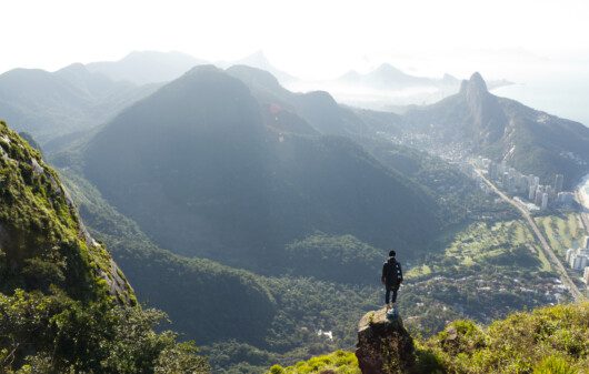 Imagem panorâmica de um homem de costas em cima da Pedra da Gávea olhando as montanhas do Rio de Janeiro para ilustrar matéria sobre parques no Rio de Janeiro