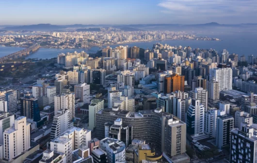 Imagem de edifícios do centro de Florianópolis vistos do alto com ponte suspensa a cabo ao fundo para ilustrar matéria sobre as melhores cidades de Santa Catarina