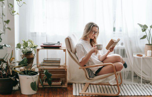 Imagem de uma mulher em uma poltrona lendo um livro e tomando café na sala da sua casa.