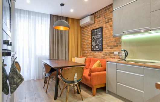 Foto de um exemplo de apartamento pequeno decorado. A imagem mostra um ambiente que integra cozinha com sala de estar e sala de jantar. Nela há um fogão embutido na parede, uma mesa com duas cadeiras, um sofá laranja e um armário de cozinha com pia embutida.