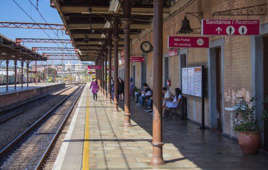 Foto que ilustra matéria sobre a Estação Jundiaí mostra a plataforma da estação de trem em um dia de céu azul, com pessoas sentadas aguardando a chegada do veículo à direita da imagem e os trilhos vazios à esquerda.