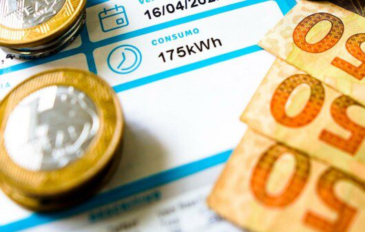 Foto que ilustra matéria sobre consumo de energia mostra uma conta de energia elétrica com moedas e notas em cima.