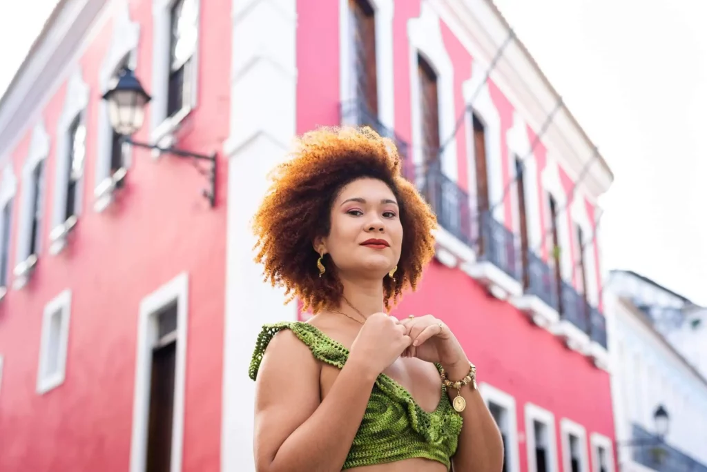 Imagem de uma mulher com cabelos ruivos sorridente em frente a um prédio colorido na cor vermelha para ilustrar matéria sobre os 5 bairros mais populosos de Salvador
