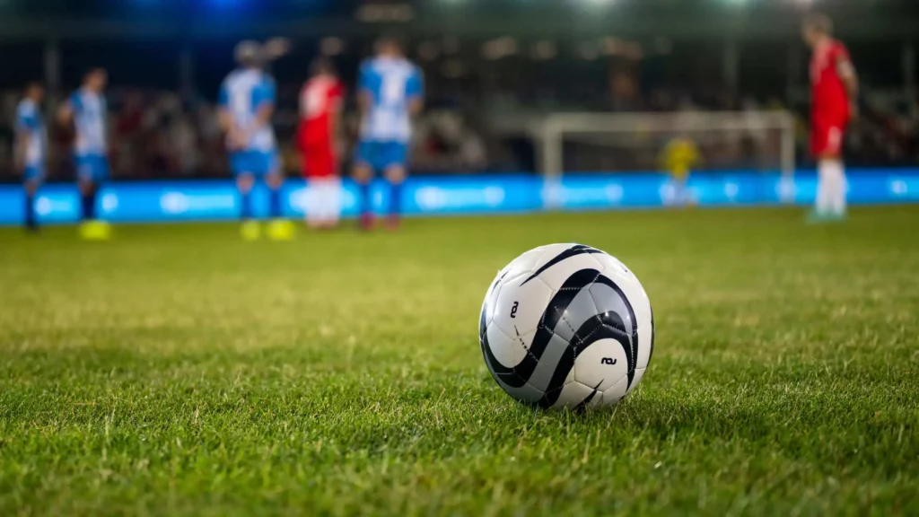 Imagem de uma bola em um gramado de futebol com vários jogadores para ilustrar matéria sobre estádios de futebol em São Paulo