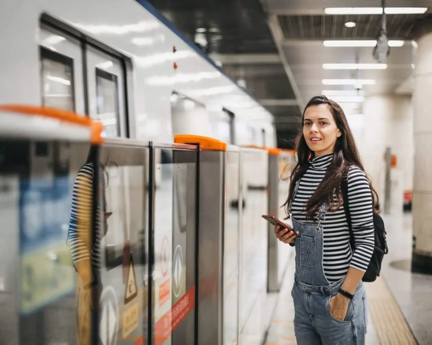 Imagem de uma mulher jovem sorrindo com o celular na mão enquanto aguarda o trem em uma estação de metrô para ilustrar matéria sobre Linha 9-Esmeralda