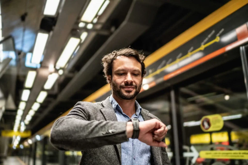 Imagem de um homem bem vestido com camiseta social e blazer olhando para o relógio em uma estação de metrô para ilustrar matéria sobre as estações da Linha Verde do metrô de São Paulo