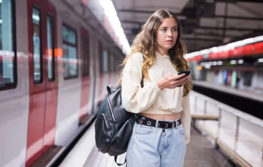 Imagem de uma mulher em uma estação de metrô com um celular na mão na frente de um trem para ilustrar matéria sobre a Linha 3-Vermelha do metrô de SP