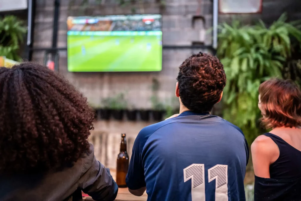 Imagem de três pessoas sentadas assistindo a um jogo de futebol na televisão para ilustrar matéria sobre qual é o maior estádio do interior paulista