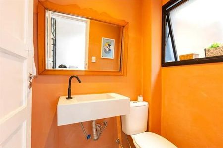 Casa à venda com 168m², 3 quartos e 1 vagaBanheiro com pia e vaso sanitário brancos, espelho e porta branca, paredes laranjas, iluminação boa, com um quadro na parede e um cesto de plantas na janela.