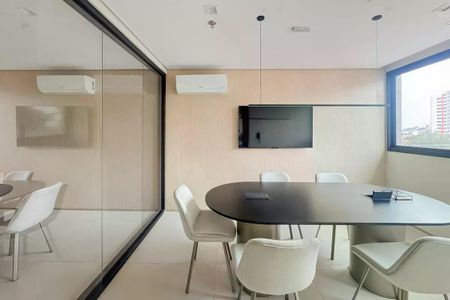 Apartamento à venda com 28m², 1 quarto e sem vagaSala de reuniões com mesa redonda e cadeiras brancas, TV na parede e janela com vista para a cidade.