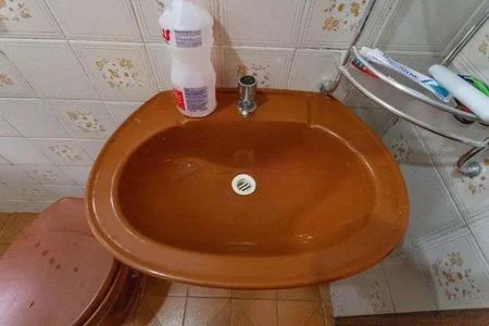 Casa à venda com 160m², 3 quartos e 2 vagasBanheiro com pia e vaso sanitário em tons de marrom, com uma garrafa de desinfetante e uma prateleira com produtos de higiene.
